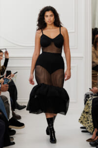 Model walking a runway in a black mesh dress with structured bodice and fishnet legs, paired with shiny black ankle boots in a white-wall setting.