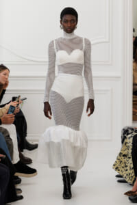 Model walks a white runway in a white mesh, full-length dress with high neck and a voluminous, ruffled hem; black patent boots with white lettering are visible.