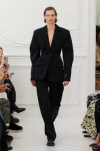 Model walks a fashion show wearing a tailored black blazer with oversized shoulders and matching black trousers.