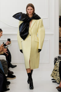 Model walks the runway in a pale yellow oversized blazer dress with a large black scarf tied around her neck and black gloves.