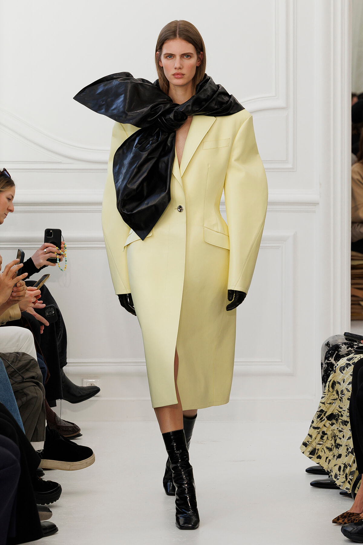 Model walks the runway in a pale yellow oversized blazer dress with a large black scarf tied around her neck and black gloves.