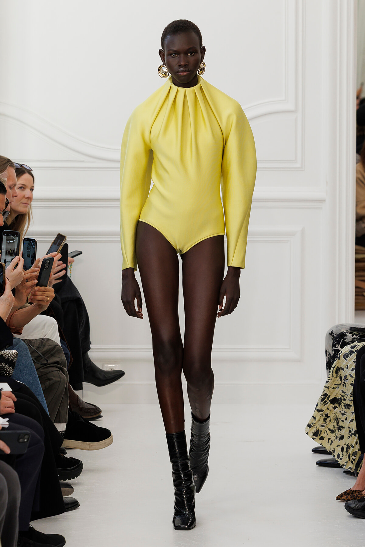 Model in a bright yellow high-neck bodysuit with long sleeves and black knee-high boots walks the runway as onlookers film with their phones.