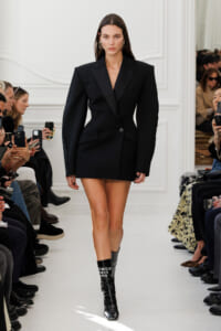 Model on a runway wearing an oversized black blazer styled as a mini dress, paired with knee-high glossy black boots with white lettering. Audience members watch and photograph the look.