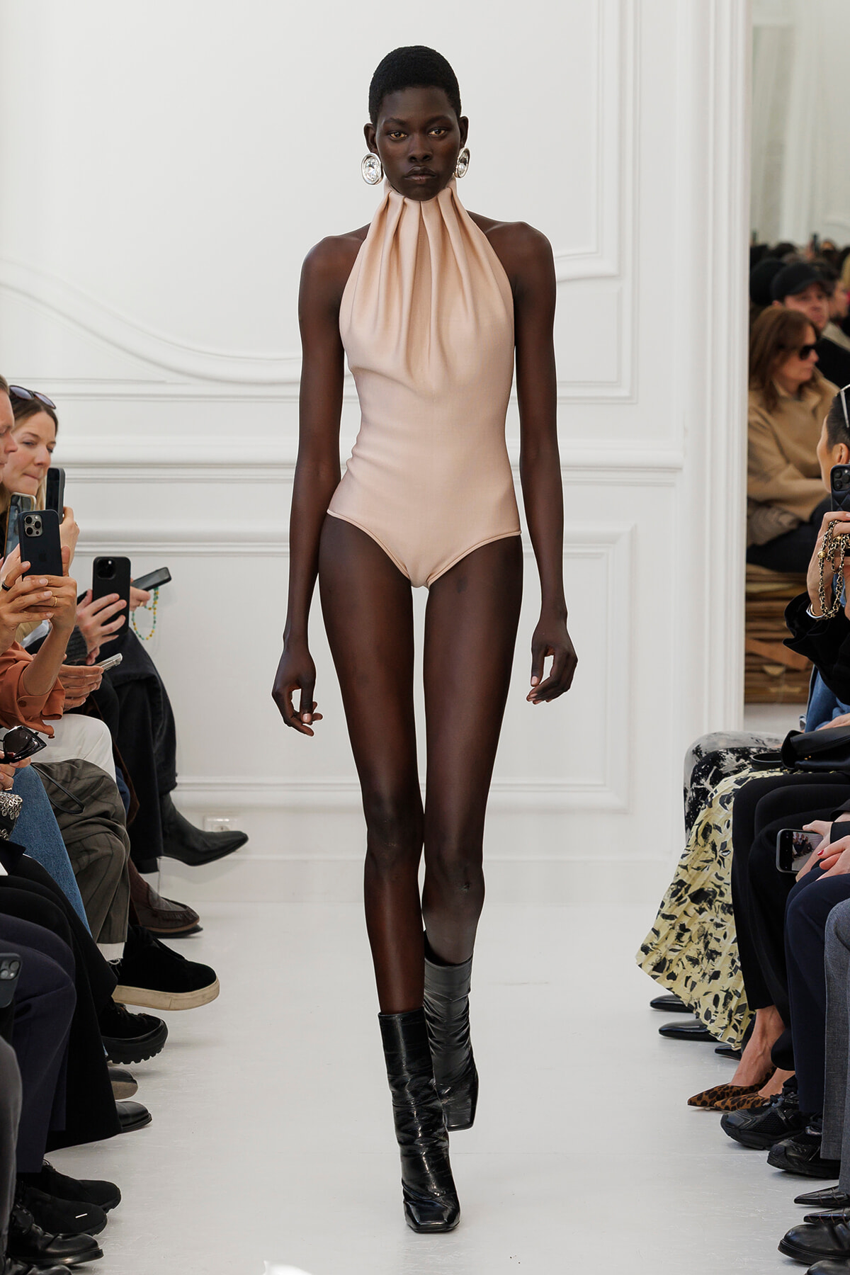 Model with dark skin walks runway in pale blush halter-neck bodysuit and black shiny boots, audience on both sides.