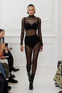 Model walks the runway wearing a black fishnet mesh bodysuit with a built‑in bra, text reading 'GIVENCHY PARIS 1952' on the chest, paired with black boots. Audience members in the foreground watch and film.