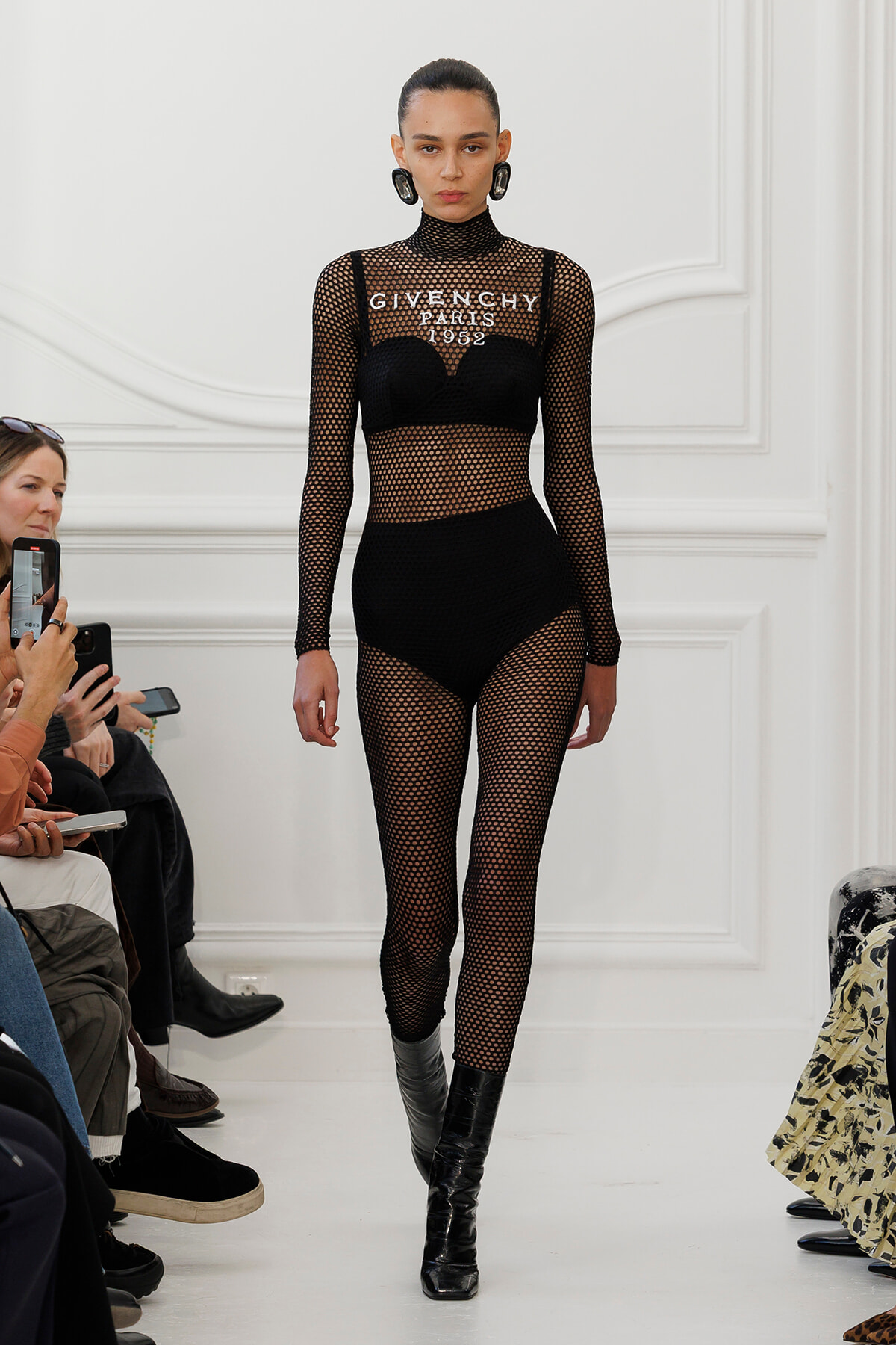 Model walks the runway wearing a black fishnet mesh bodysuit with a built‑in bra, text reading 'GIVENCHY PARIS 1952' on the chest, paired with black boots. Audience members in the foreground watch and film.