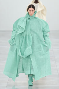Runway model wearing an oversized mint-green knitted coat with a chunky scarf and matching boots in a fashion show setting.