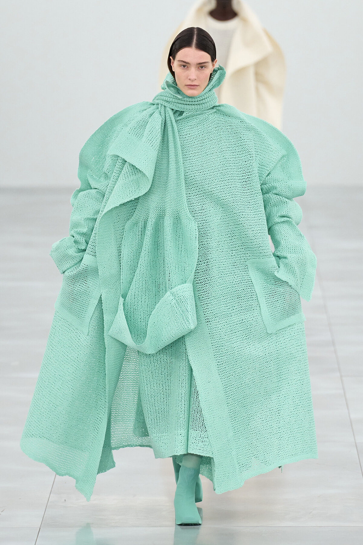 Runway model wearing an oversized mint-green knitted coat with a chunky scarf and matching boots in a fashion show setting.