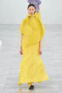 Runway model in a bright yellow pleated gown with an oversized draped wrap and high-neck scarf.