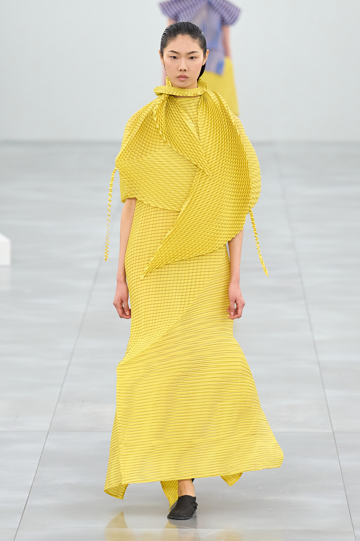 Runway model in a bright yellow pleated gown with an oversized draped wrap and high-neck scarf.