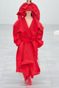 Model walks a runway in a voluminous bright red knit coat with a wide belt and matching hat.