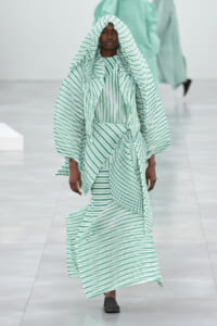 Model walking a fashion runway in a mint green and white striped oversized hooded cape and matching dress set.