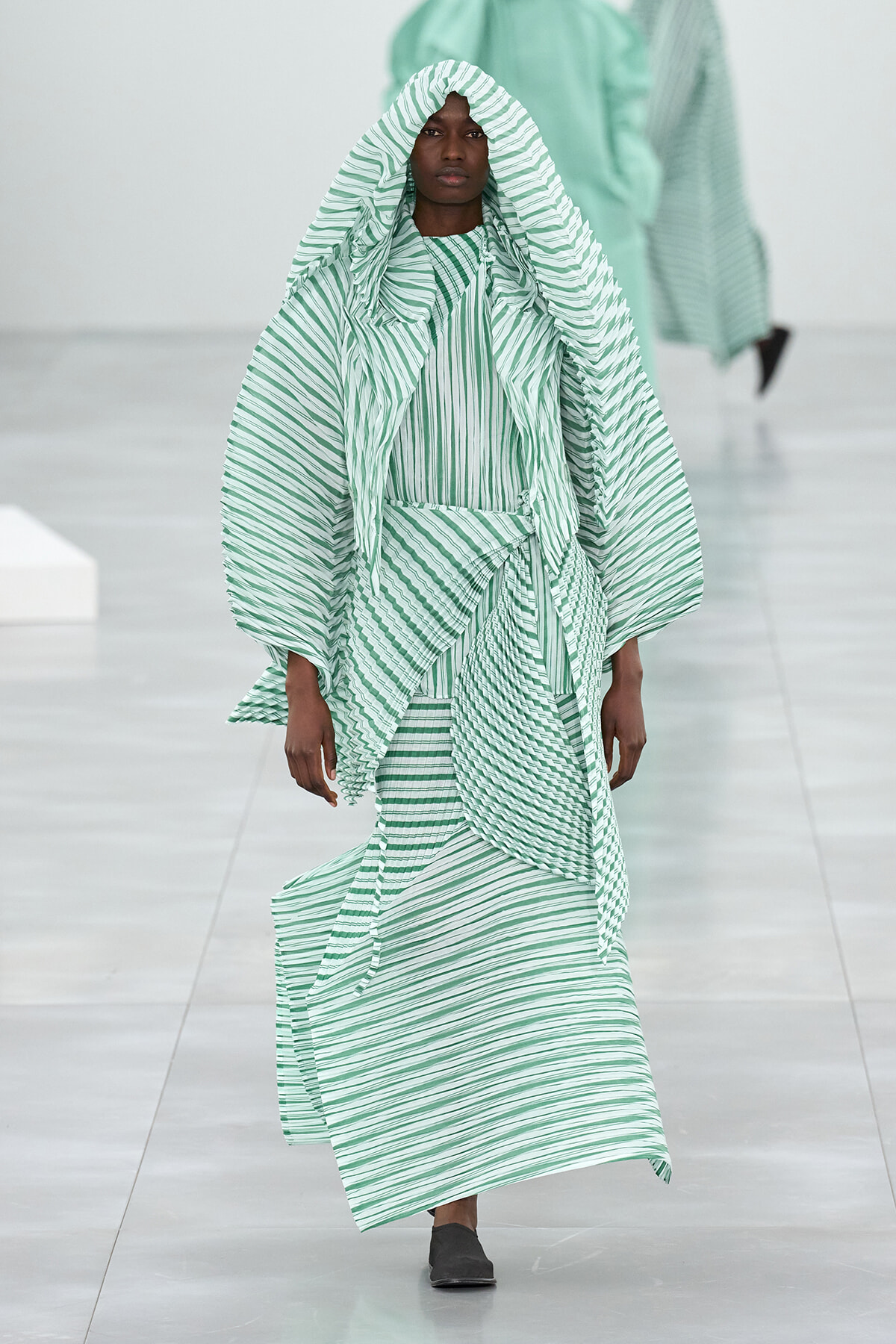 Model walking a fashion runway in a mint green and white striped oversized hooded cape and matching dress set.
