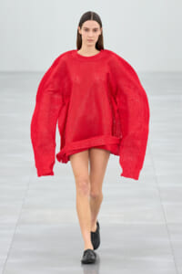 Model walking a runway in a bright red oversized knitted sweater with balloon sleeves and a short hem, paired with black slip-on shoes.