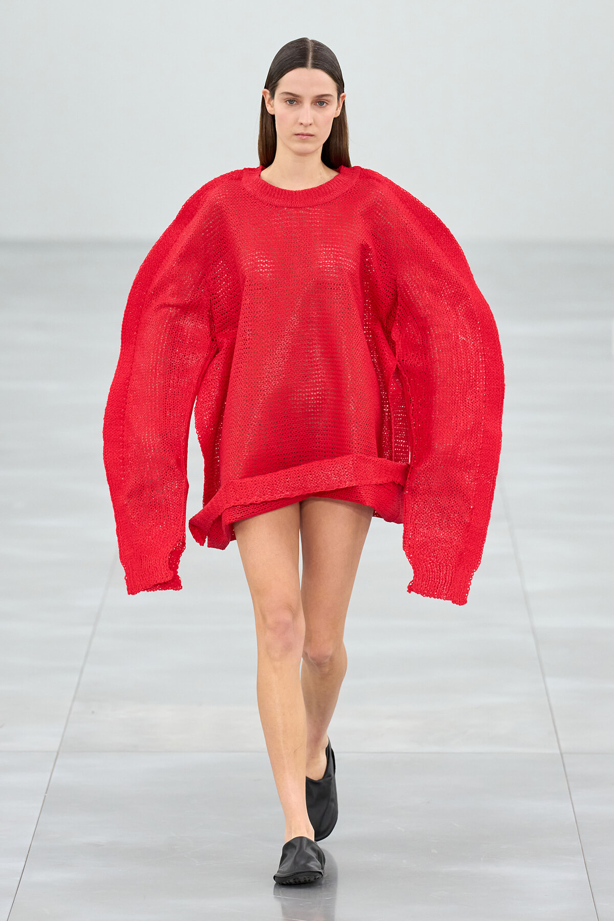 Model walking a runway in a bright red oversized knitted sweater with balloon sleeves and a short hem, paired with black slip-on shoes.