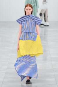 Model walking a runway in a blue and white striped top with wide flutter sleeves and a bright yellow pleated skirt, black shoes.