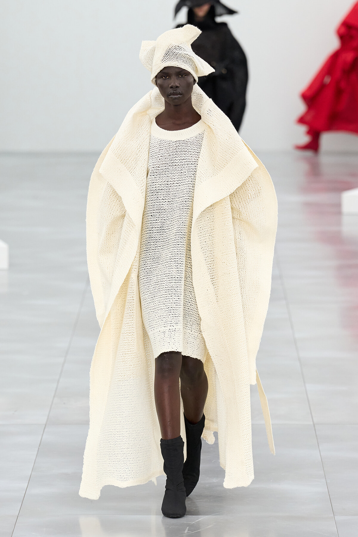Model walks the runway in an oversized cream knit cape over a matching dress and a headwrap, black ankle boots visible.