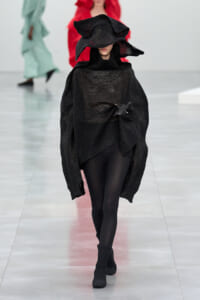 Model on a fashion runway wearing a black oversized knit poncho with a dramatic sculptural hood and black tights.
