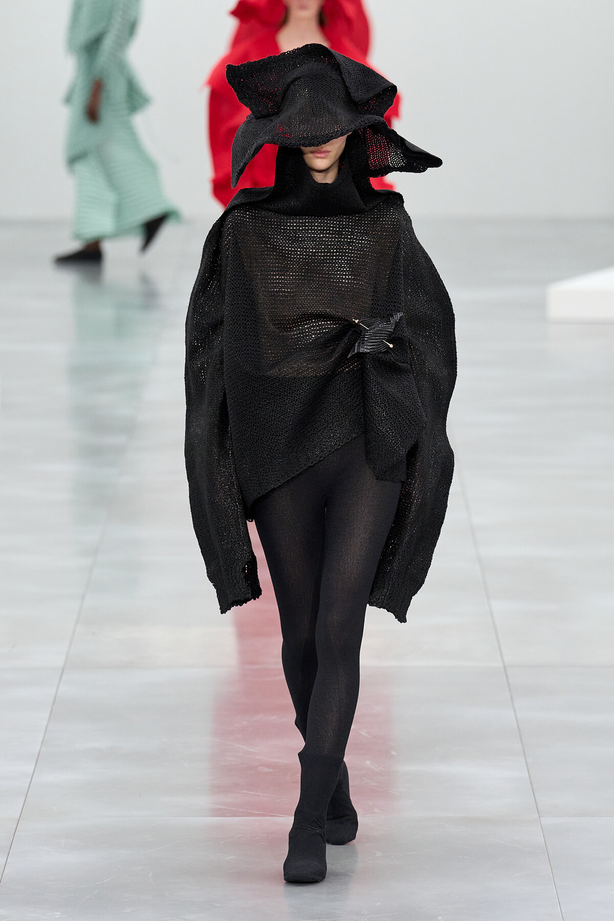 Model on a fashion runway wearing a black oversized knit poncho with a dramatic sculptural hood and black tights.
