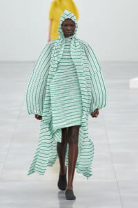 Model walking a runway in a green and white striped oversized cape and matching hooded dress.