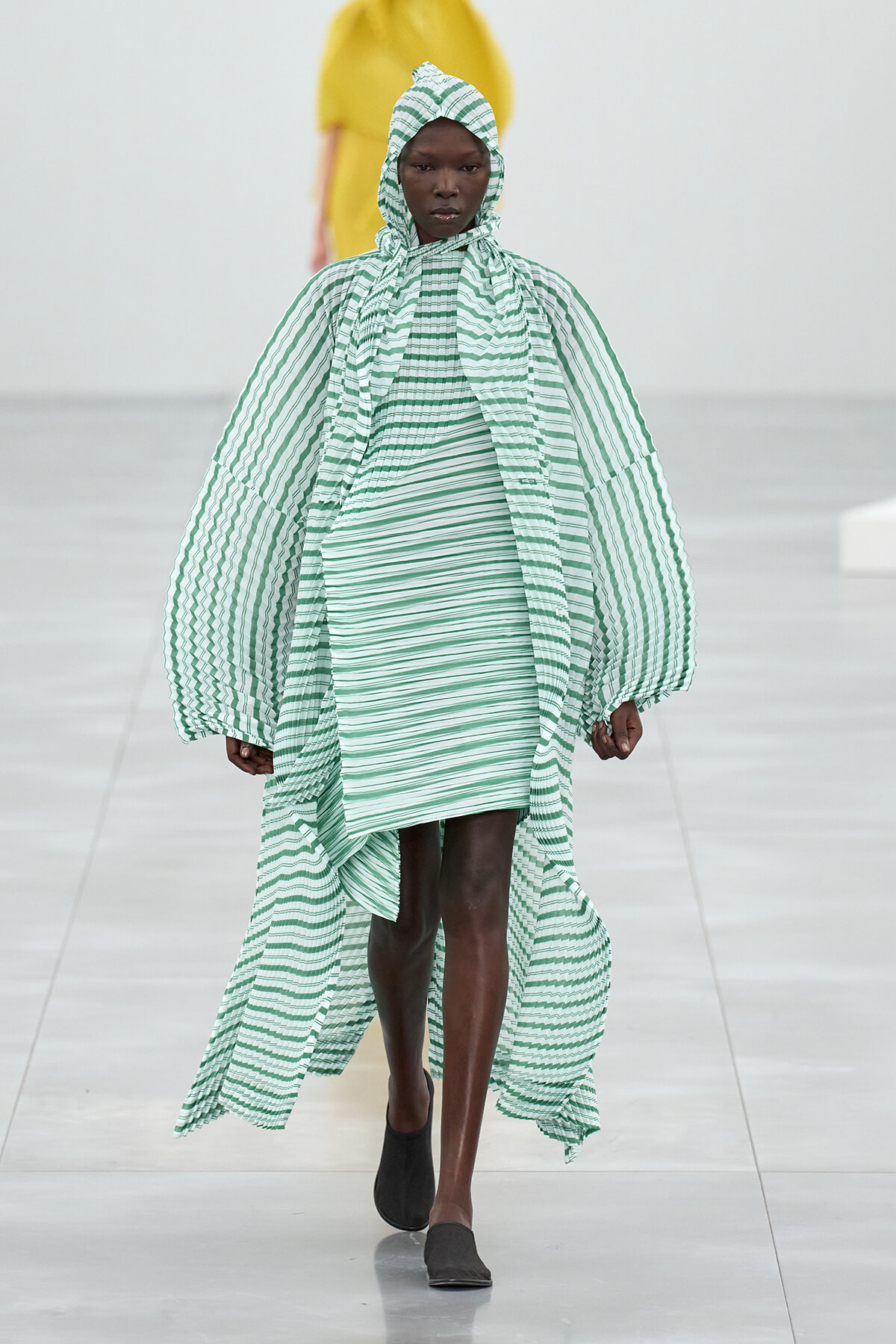 Model walking a runway in a green and white striped oversized cape and matching hooded dress.