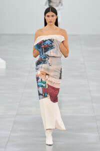 Model walking the runway in an off‑the‑shoulder printed maxi dress with abstract blue, beige, and red patterns, paired with white boots.