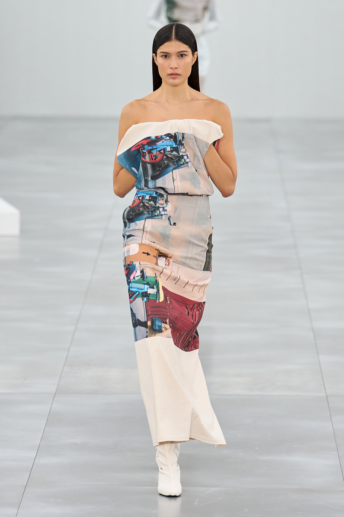 Model walking the runway in an off‑the‑shoulder printed maxi dress with abstract blue, beige, and red patterns, paired with white boots.
