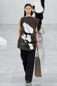 Model walking a runway in an oversized olive top with white abstract shapes and black wide-leg pants, carrying a brown tote bag.