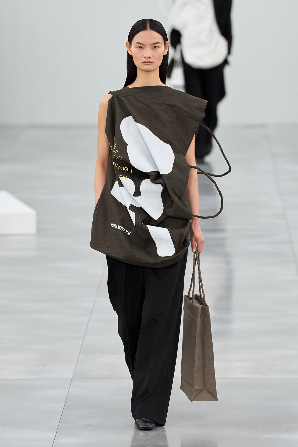 Model walking a runway in an oversized olive top with white abstract shapes and black wide-leg pants, carrying a brown tote bag.