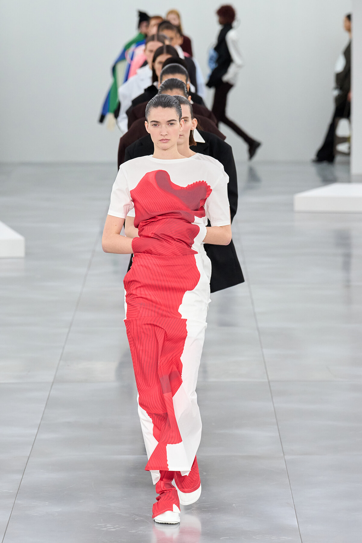 Front view of a female model walking the runway in a red and white color-block dress with a bold abstract print, others lined up behind her.