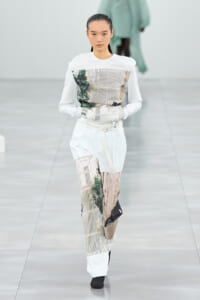 Female model on a runway wearing a white top with graphic printed panels and light patchwork pants with a belt.