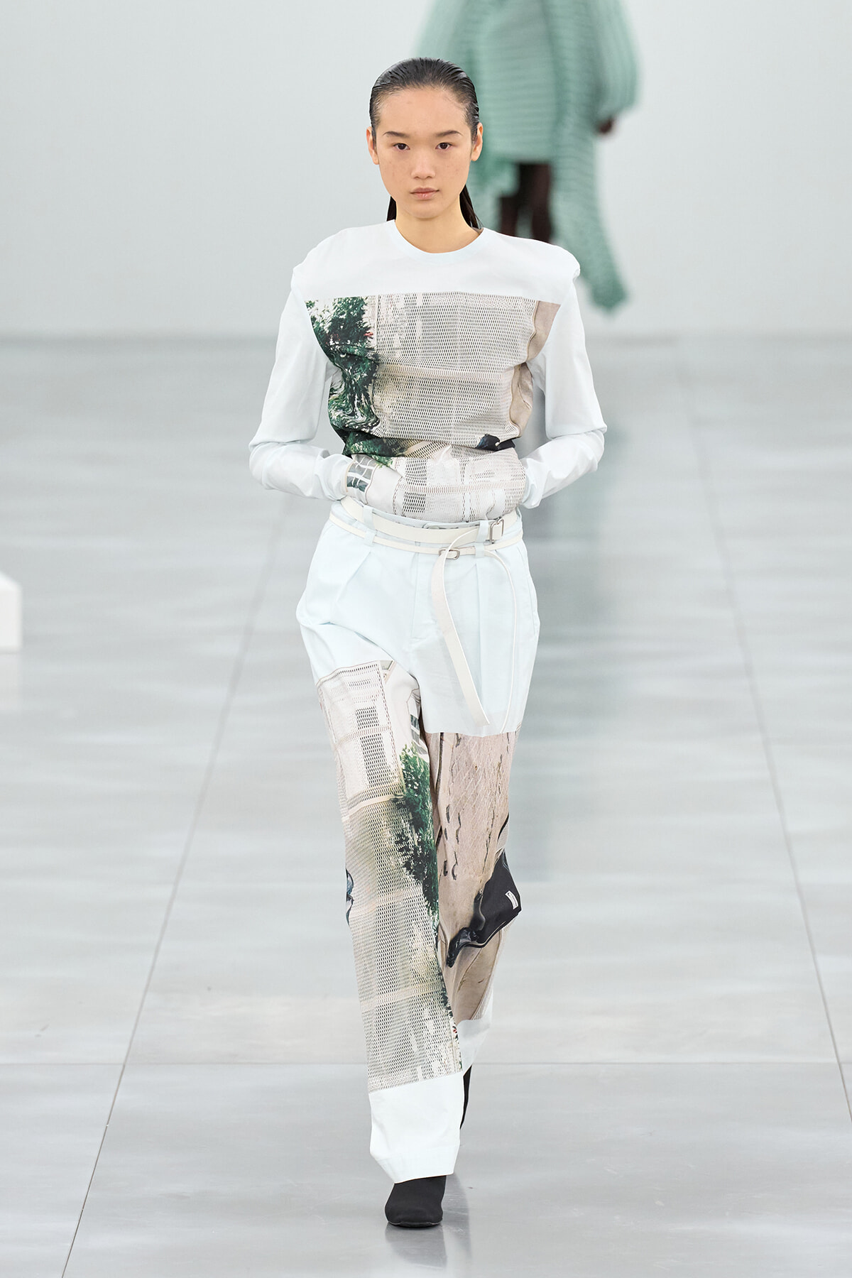 Female model on a runway wearing a white top with graphic printed panels and light patchwork pants with a belt.