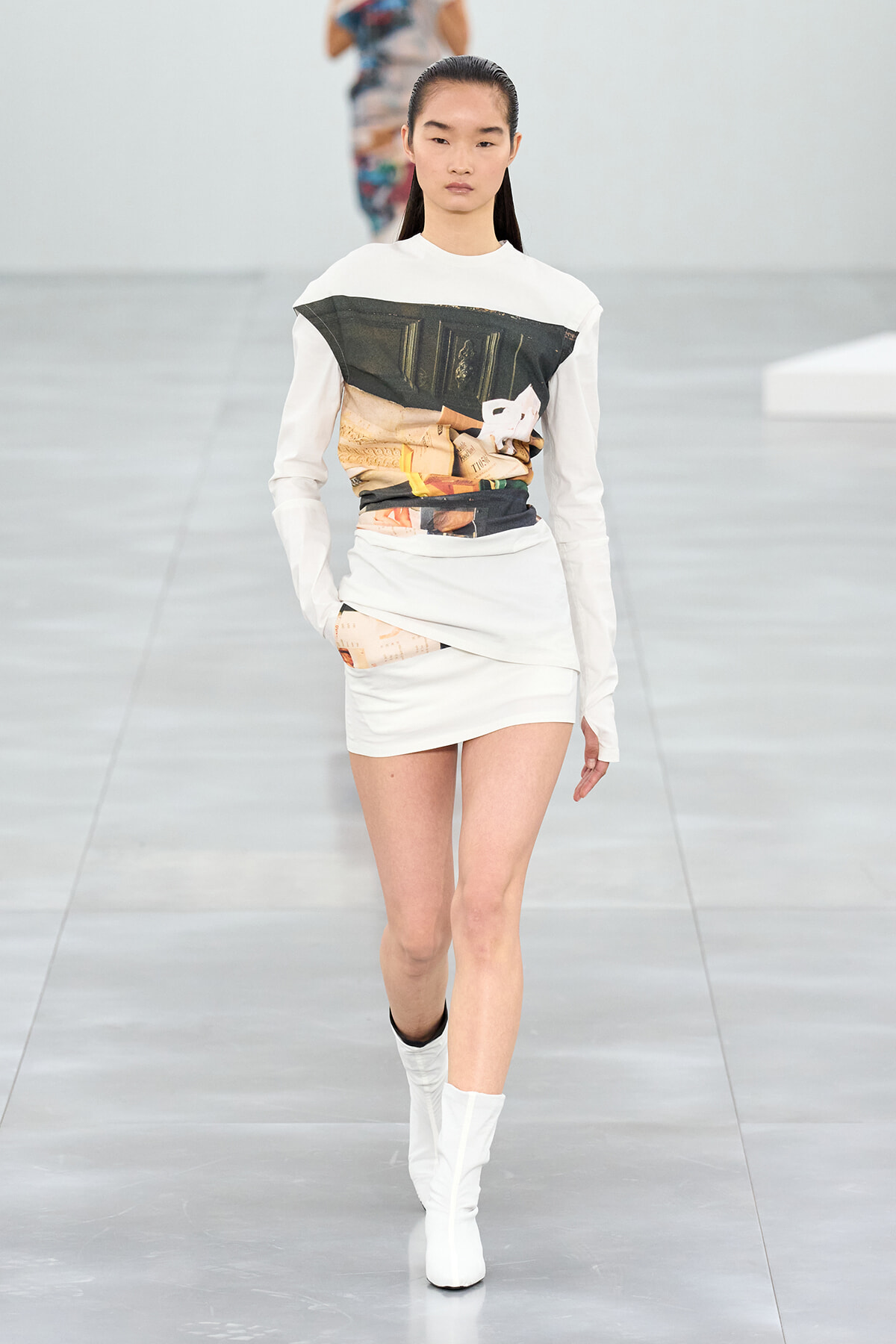 Model walking the runway in a white long-sleeve top with a large abstract graphic panel and a white, wrapped mini skirt with a printed accent, paired with white ankle boots.