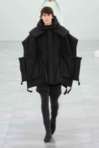 Model walks in a loose black puffer poncho with oversized collar and large pocketed panels on a runway.