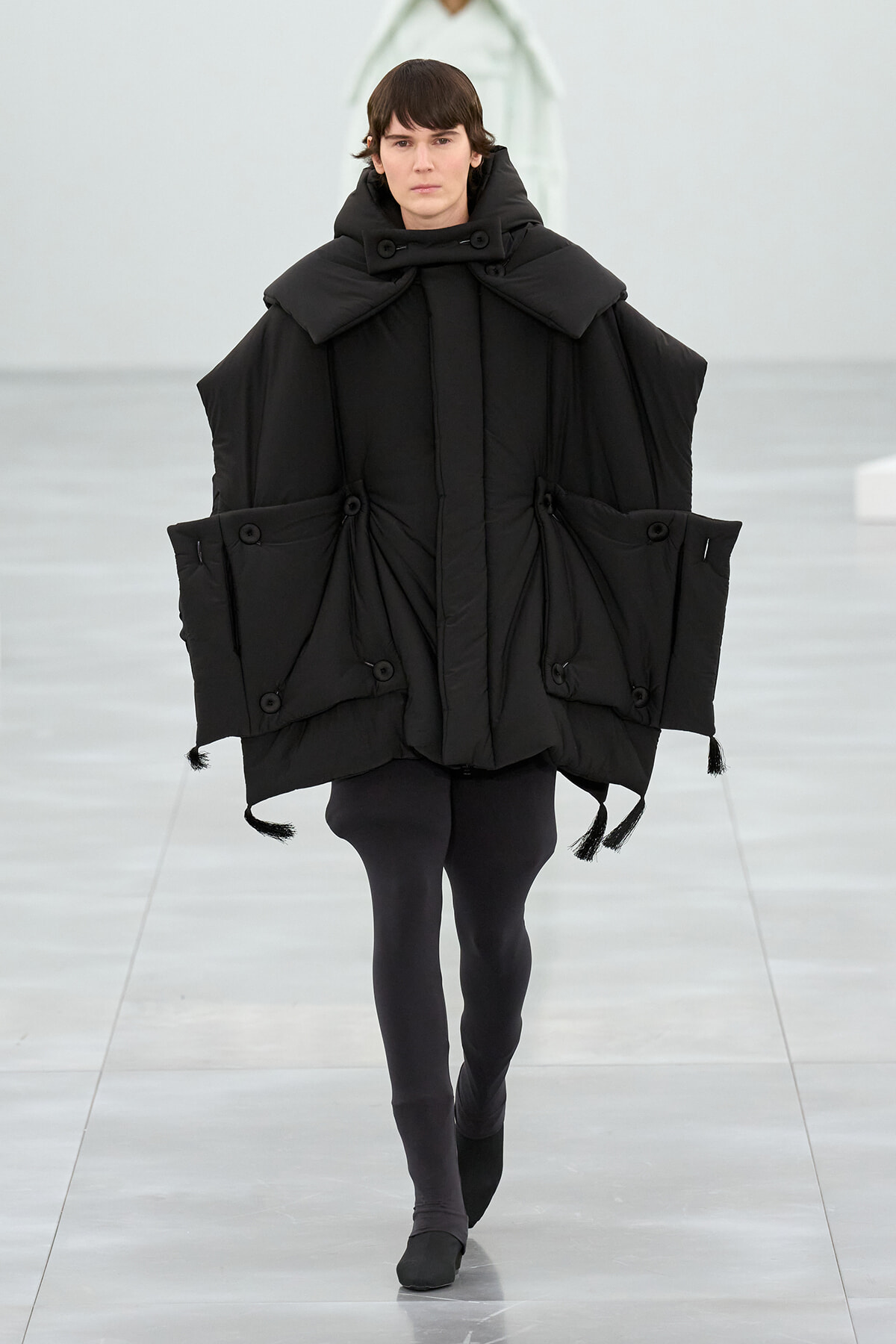 Model walks in a loose black puffer poncho with oversized collar and large pocketed panels on a runway.