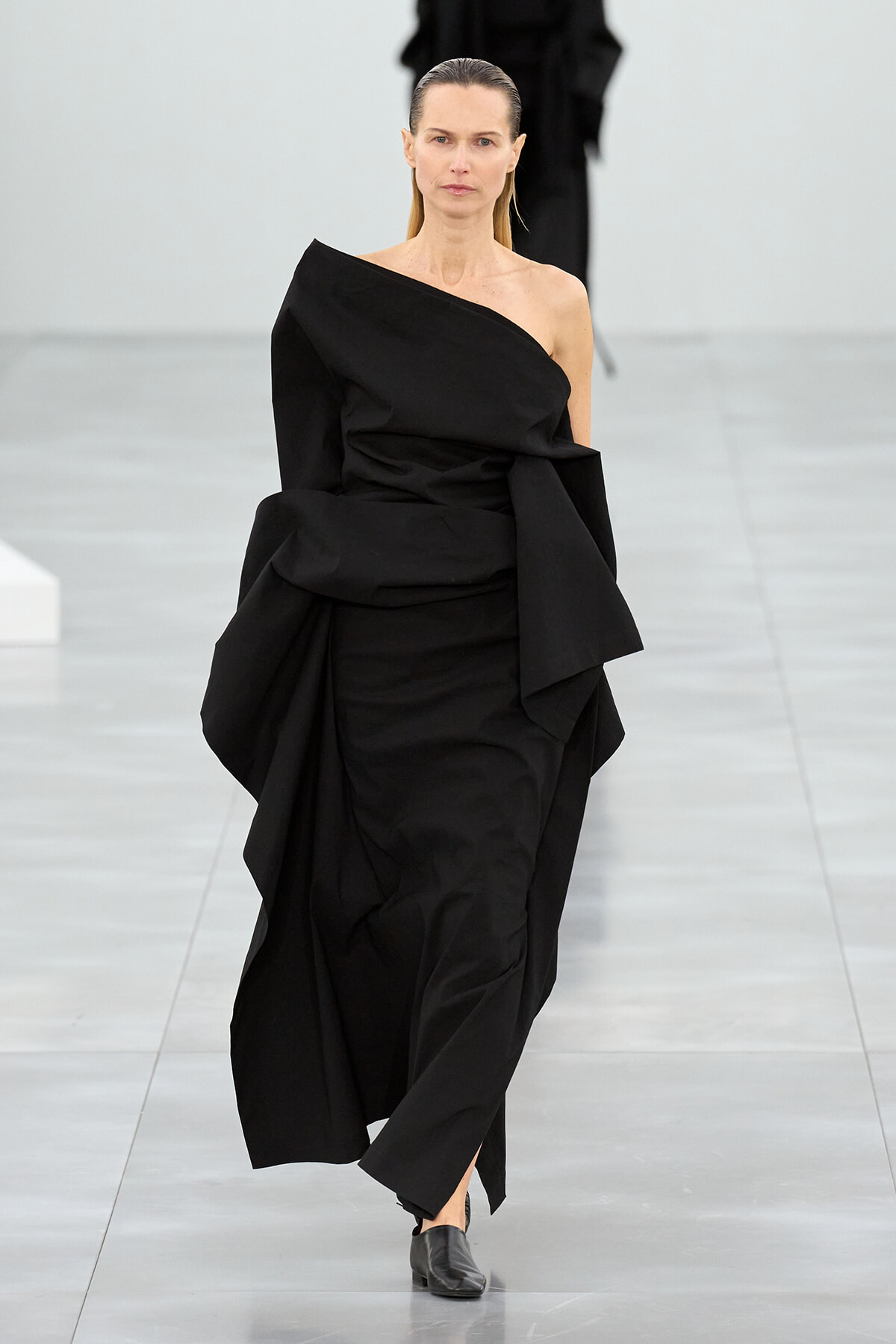 Model walking a fashion runway in a black one-shoulder, draped gown with sculptural folds.