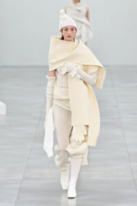 Model walks the runway in a cream-toned avant-garde knit outfit with scarf, gloves, and turban-style head wrap on a pale runway.