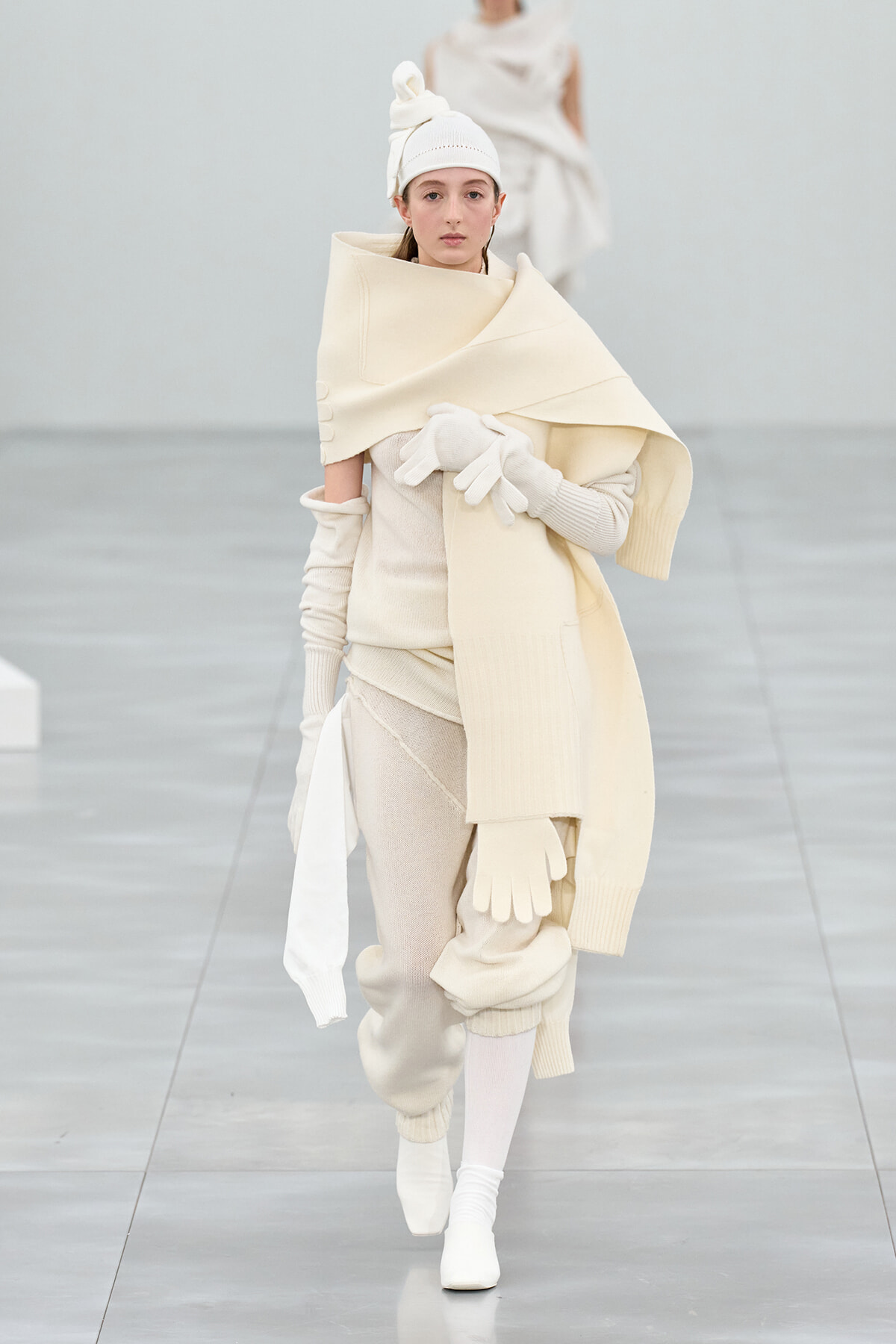 Model walks the runway in a cream-toned avant-garde knit outfit with scarf, gloves, and turban-style head wrap on a pale runway.
