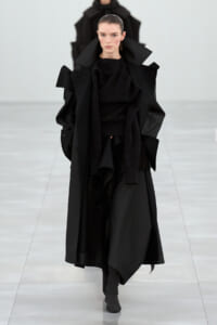 Model wearing an oversized black coat with dramatic lapels and a layered scarf on a runway runway show