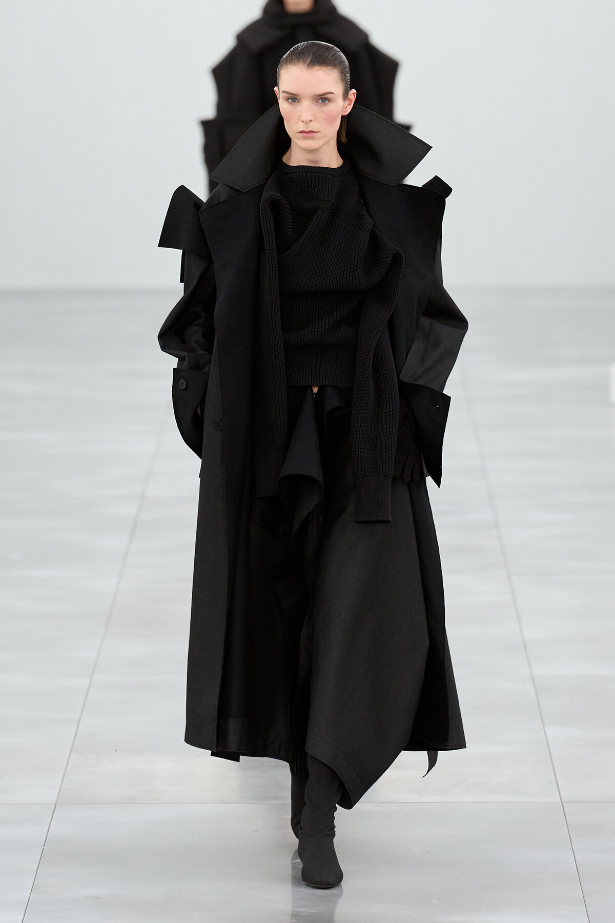 Model wearing an oversized black coat with dramatic lapels and a layered scarf on a runway runway show
