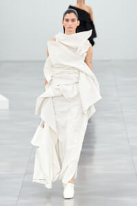 Model walking a runway in a sculptural white draped gown with oversized folds and a high collar.