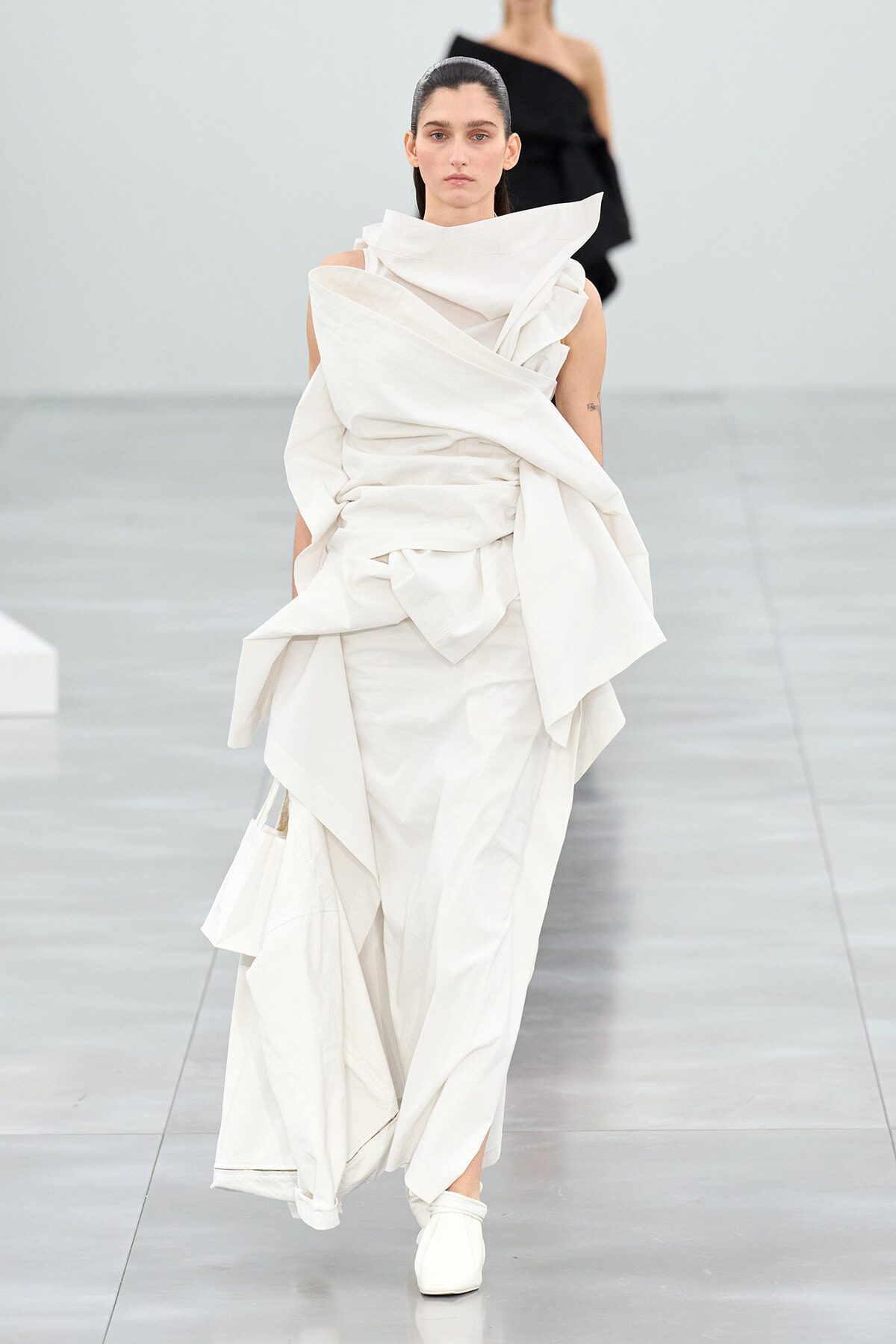 Model walking a runway in a sculptural white draped gown with oversized folds and a high collar.