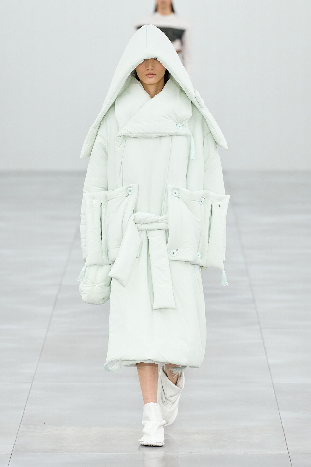 Model walking a runway in a voluminous pale mint-green hooded puffer coat with a belt and oversized pockets.
