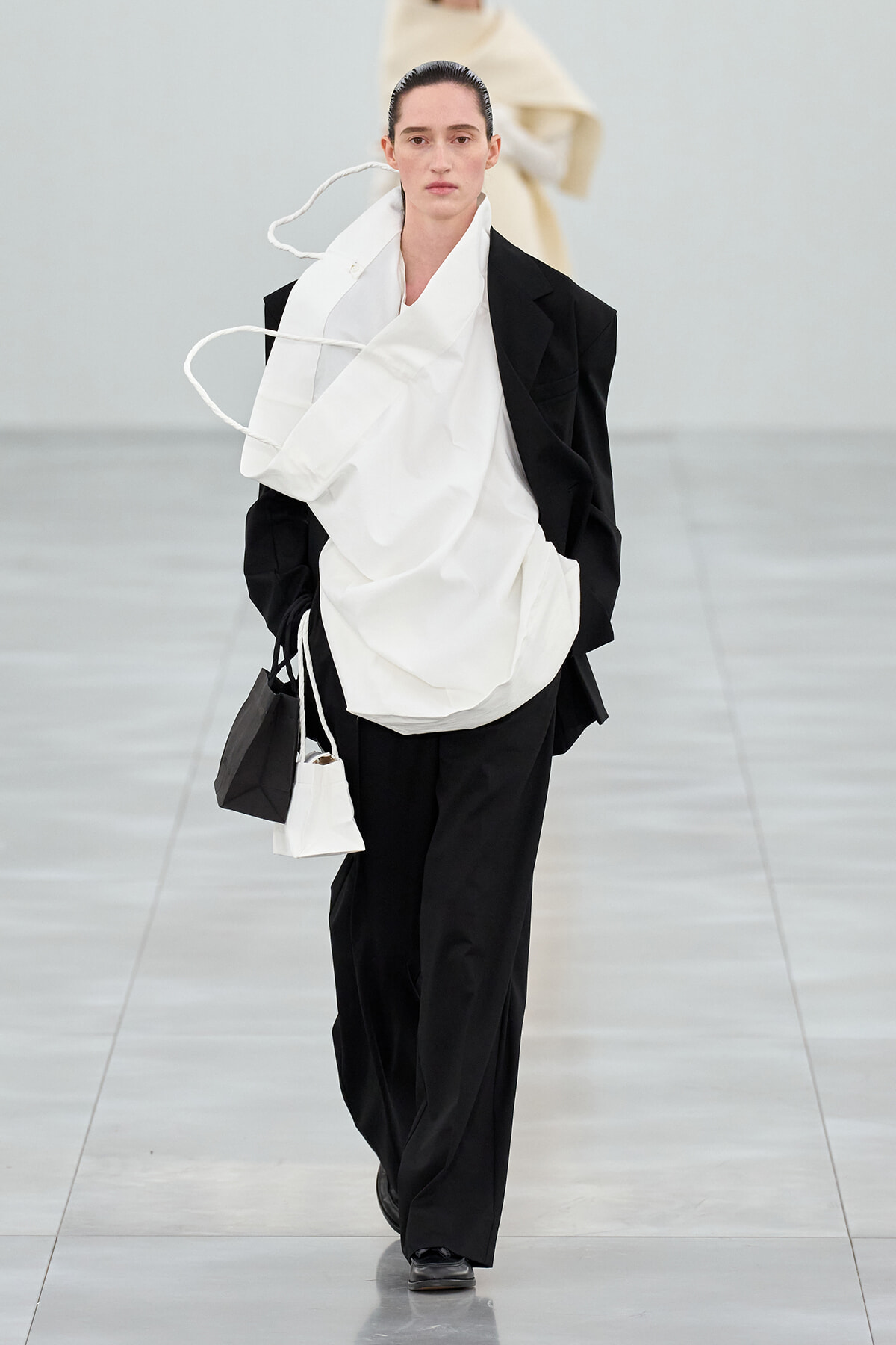 Model walking a runway in an oversized black suit with a large white wrap, carrying black and white handbags with rope accents.