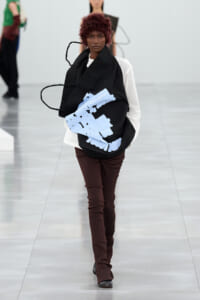 Model walking a runway with a large black tote featuring light-blue abstract shapes and the Issey Miyake logo on the bag to the front.