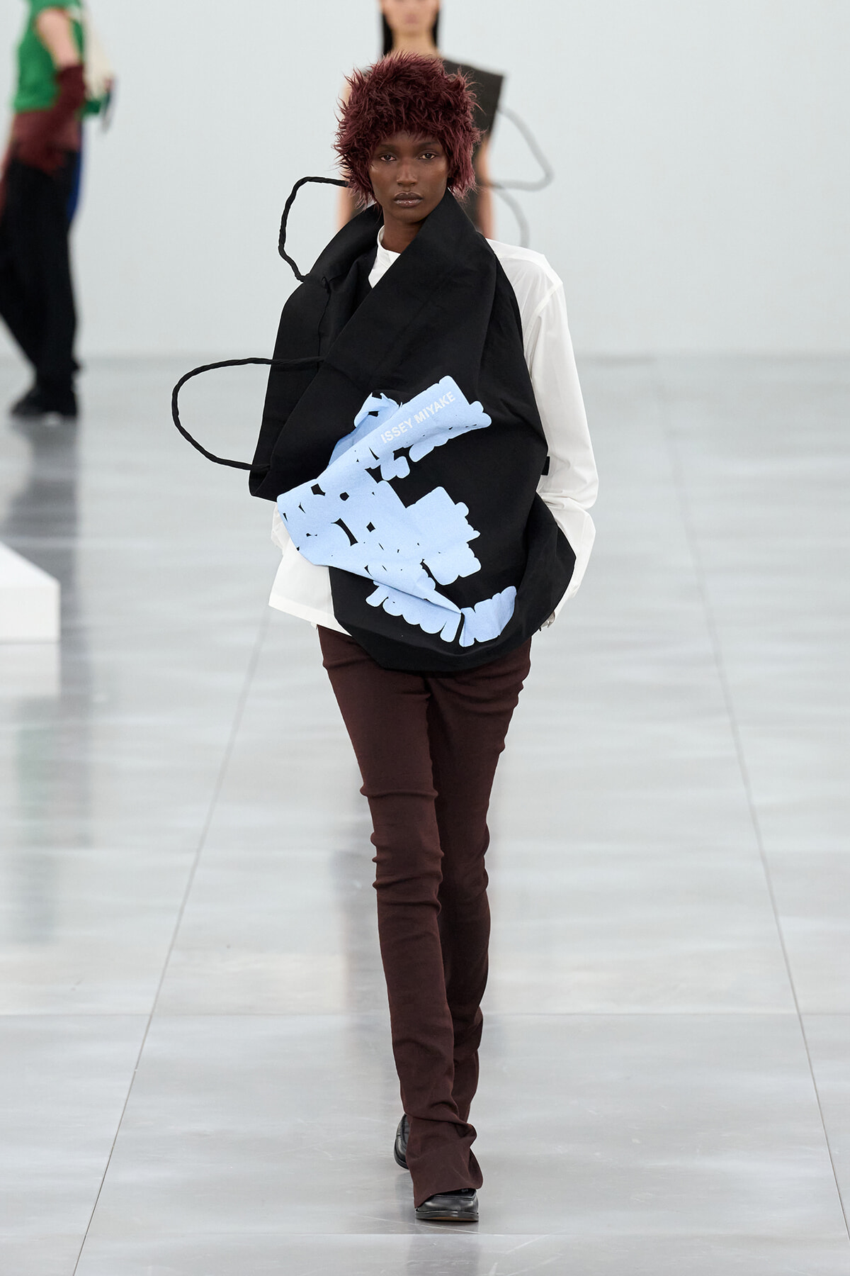 Model walking a runway with a large black tote featuring light-blue abstract shapes and the Issey Miyake logo on the bag to the front.