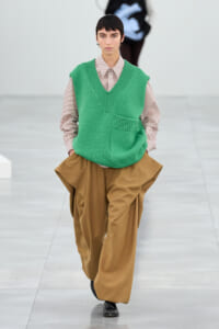 Model walks the runway wearing a bright green V-neck knit vest over a striped button-down shirt and oversized tan trousers on a minimalist catwalk.