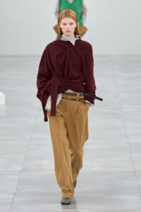 Model walking a runway in a burgundy knit sweater cinched at the waist, striped shirt peeking out, tan wide trousers with a belt.