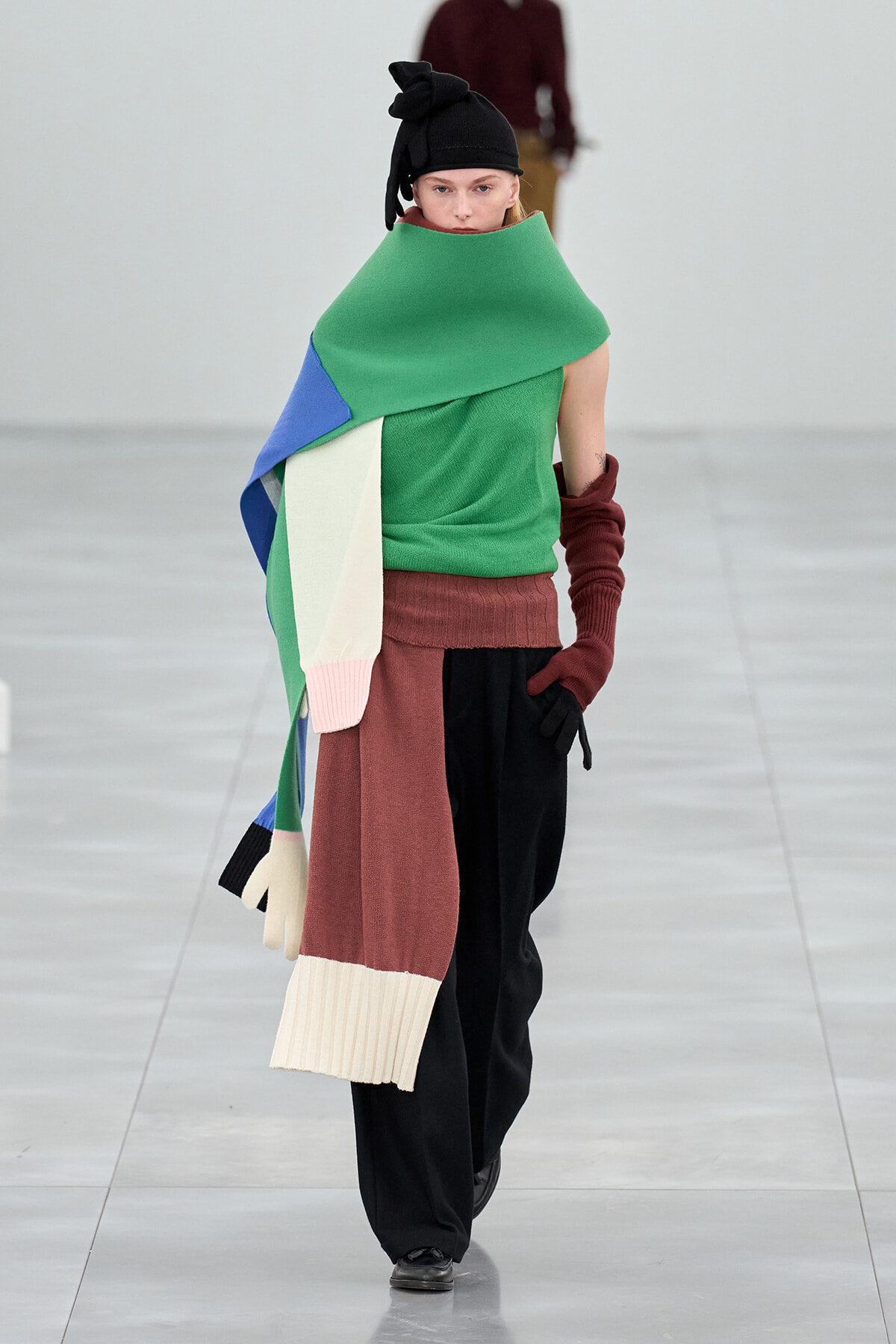 Model walks the runway in an oversized color-block scarf draped over a green top, with maroon gloves and black pants, wearing a black knot-tied hat.