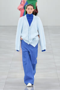 Model walks the runway in a pale blue oversized cardigan over a bright blue turtleneck and matching wide-leg pants, hands in pockets.